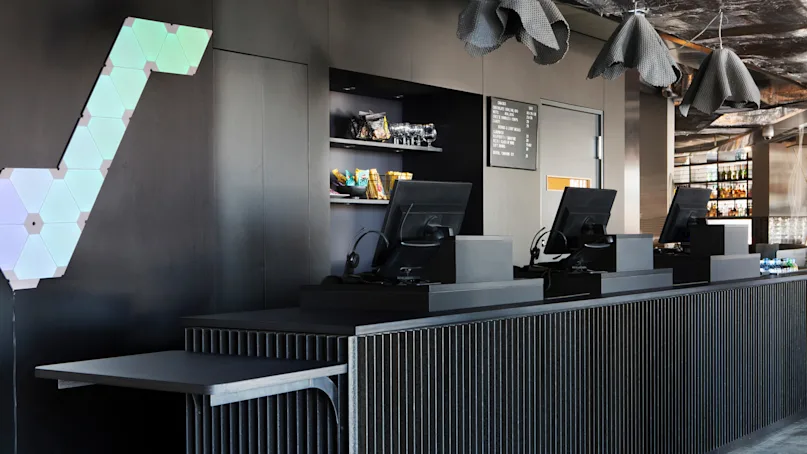 A minimalist black counter with two computer terminals, shelves of snacks in the background, and geometric lights on the wall.