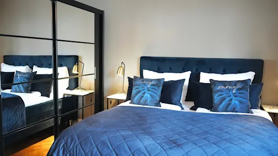 En blå seng med dekorative puter merket "enough" står mellom to gullamper, reflektert i et speilgarderobe i et moderne soverom.