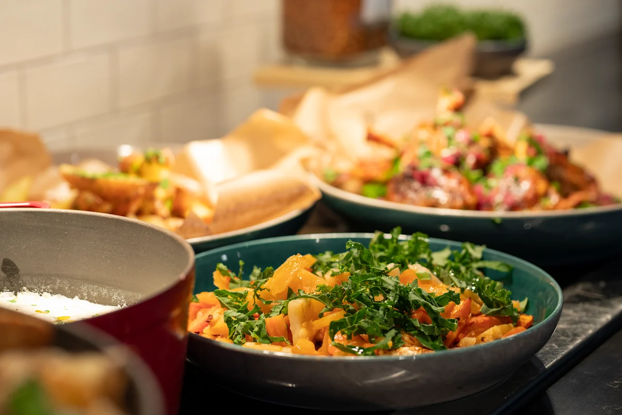 Swedish: En blå skål innehåller en livfull sallad med grönt och grönsaker, omgiven av fler tallrikar med färgglad mat på en köksbänk, under varm belysning.
