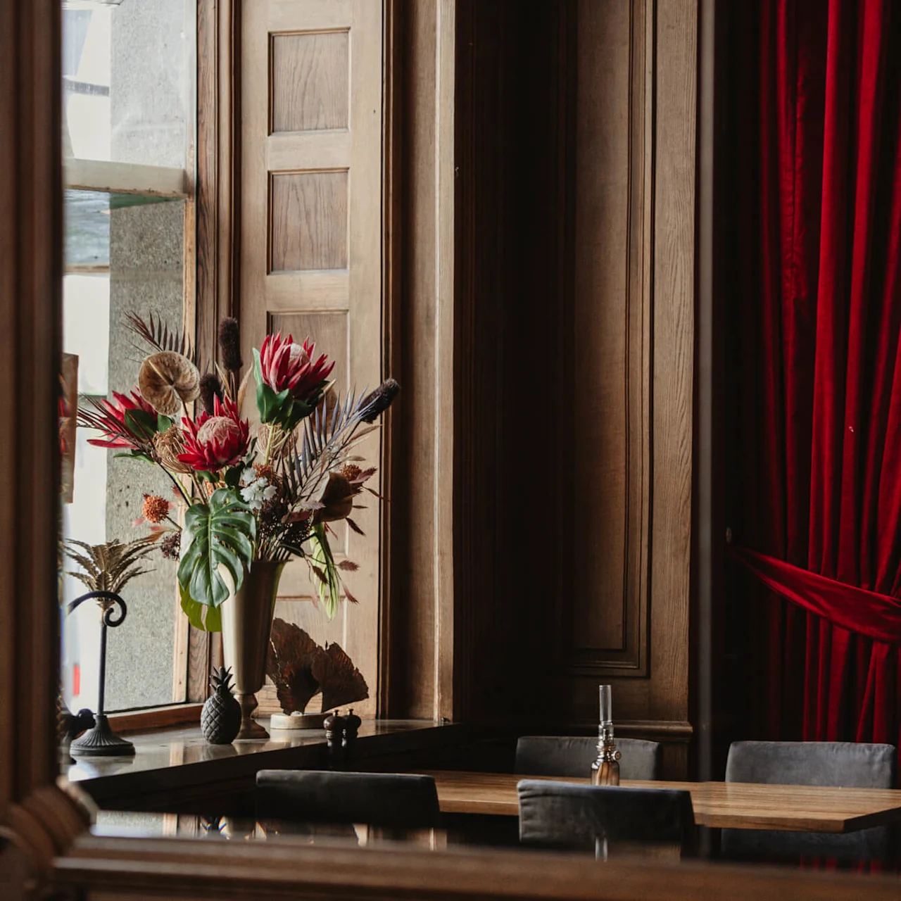 En vase med fargerike blomster står på en treoverflate, reflektert i et speil, med luksuriøse røde gardiner i et elegant rom.