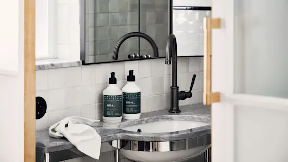 A bathroom sink with a black faucet stands in a modern setting. Two bottles labeled “SKOG” rest beside a rolled white towel on a marble countertop.