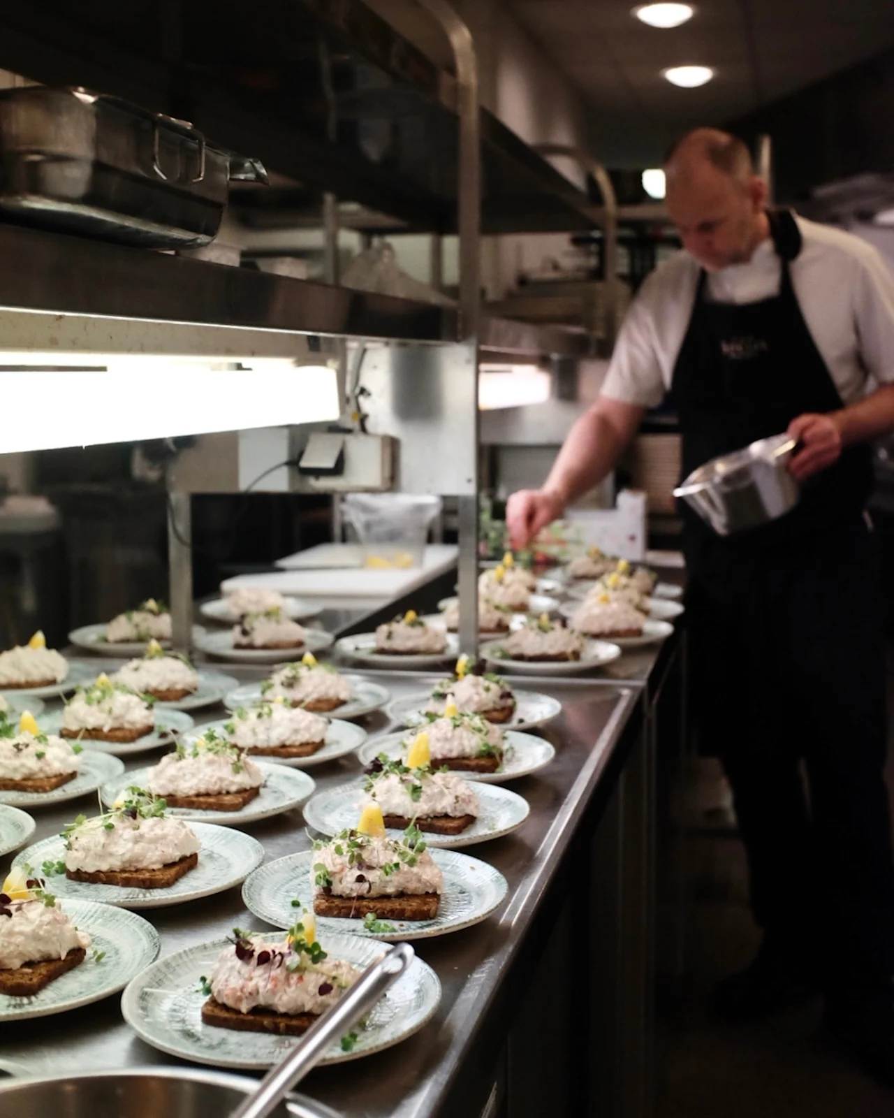 Plates with open-faced sandwiches are being garnished by a chef in a professional kitchen. The sandwiches are arranged neatly on a stainless steel counter under bright lights.