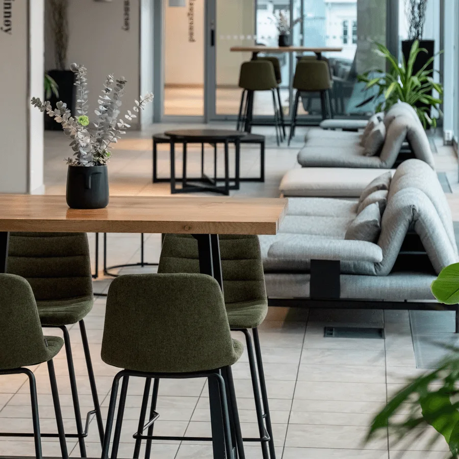 Modernt träbord med gröna stoppade stolar runt, med en svart vas med dekorativa löv; i en elegant, kaklad lounge med soffor och växter under naturligt ljus.