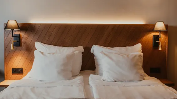 Viihtyisä hotellisänky puupäädyllä, valkoisilla tyynyillä ja lämpimällä valaistuksella yöpöytävalaisimista, kutsuen lepoon.