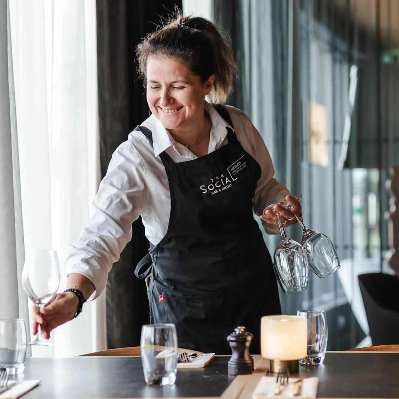 A server, wearing an apron labeled “The Social”, sets glasses on a table in a restaurant. The table is elegantly arranged with cutlery, glassware, and a lit candle.