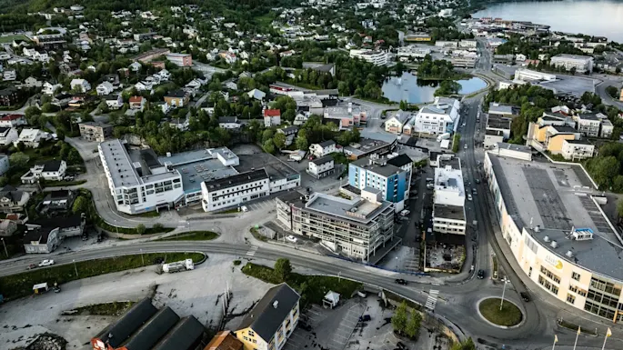 Aerial view of a small town with scattered buildings and greenery, nestled between a lake on the right and mountains in the background beneath a partly cloudy sky.