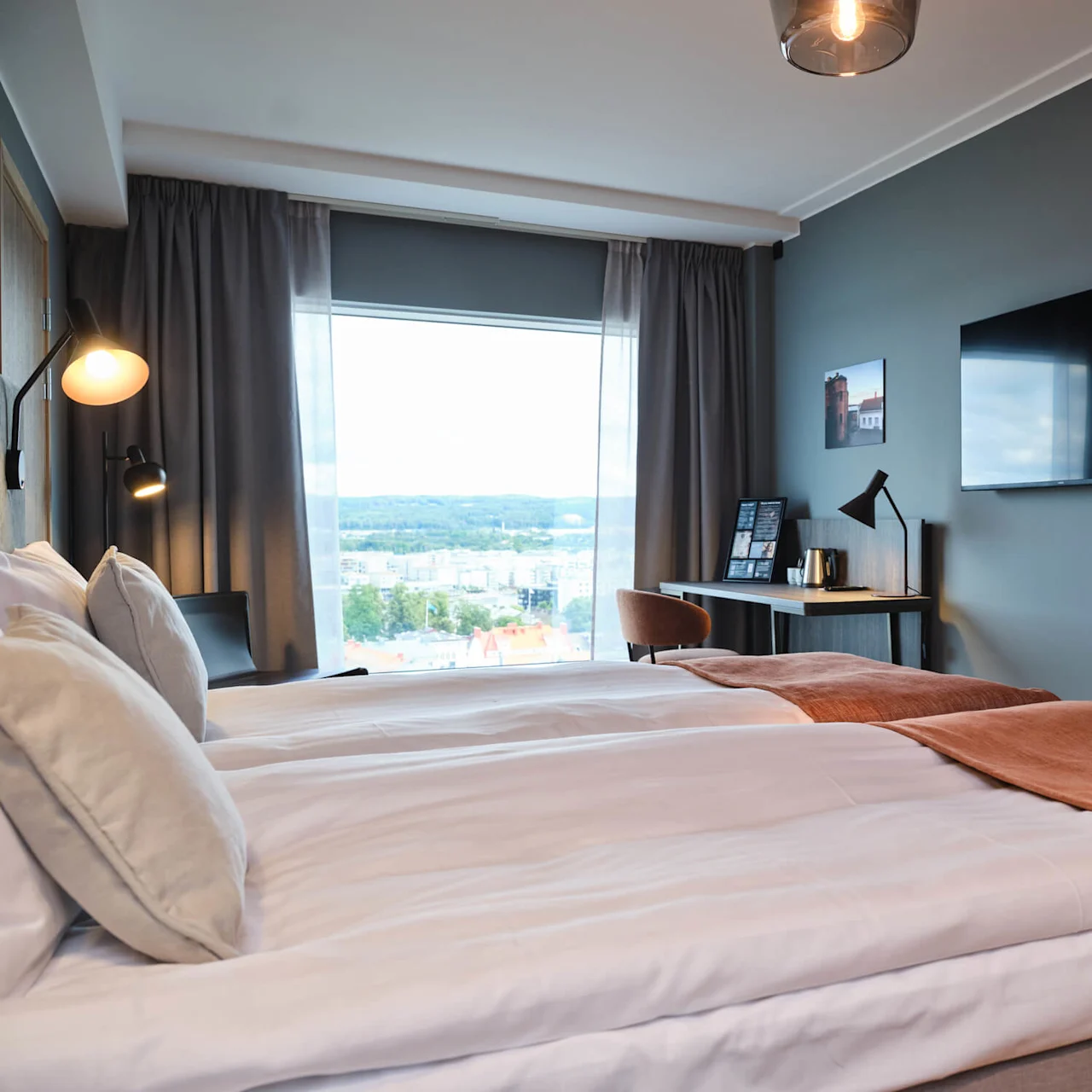 En pænt redt seng med et rustfarvet tæppe står ved siden af et vindue med udsigt over bybilledet. Et skrivebord med lampe og et TV på væggen fuldender det moderne rum.