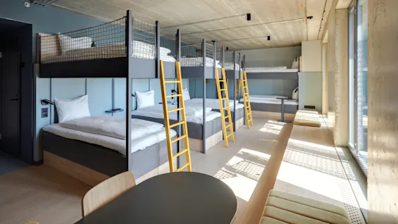 Køjesenge med gule stiger er opstillet langs en væg. Sollys strømmer ind gennem gulv-til-loft-vinduer og oplyser et moderne, minimalistisk rum med trægulve og siddepladser.