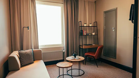Ett mysigt hotellrum med en soffa, två soffbord, ett skrivbord och en stol, badat i naturligt ljus från ett stort fönster.