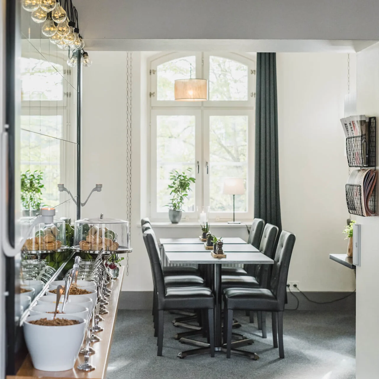 Bright dining room with a long table set for a meal, surrounded by chairs, adjacent to a food serving area, bathed in natural light from large windows.