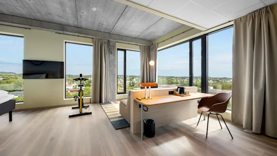 A modern hotel room with large windows, a dedicated workspace, and an exercise bike for a balanced stay.