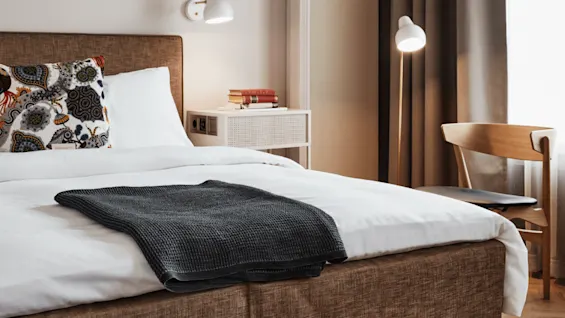 En pent oppredd seng med et mønstret putevar og mørkt teppe står i et koselig soverom, ved siden av et lite hvitt nattbord med bøker, under varm belysning ved et vindu.