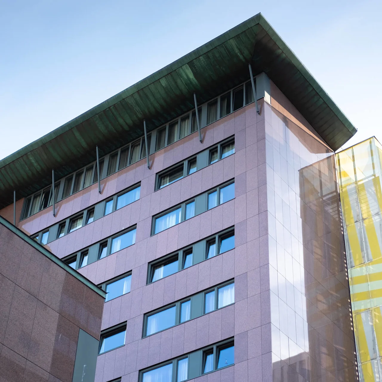 A modern multistory building with pink and glass facade, featuring rows of windows, stands under a clear blue sky. A yellow-tinted glass extension is on the right side.