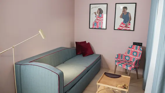 En blå soffa med vinröda kuddar står under en vit lampa, bredvid en färgglad stol. Två inramade illustrationer hänger på den rosa väggen ovanför ett litet träbord.