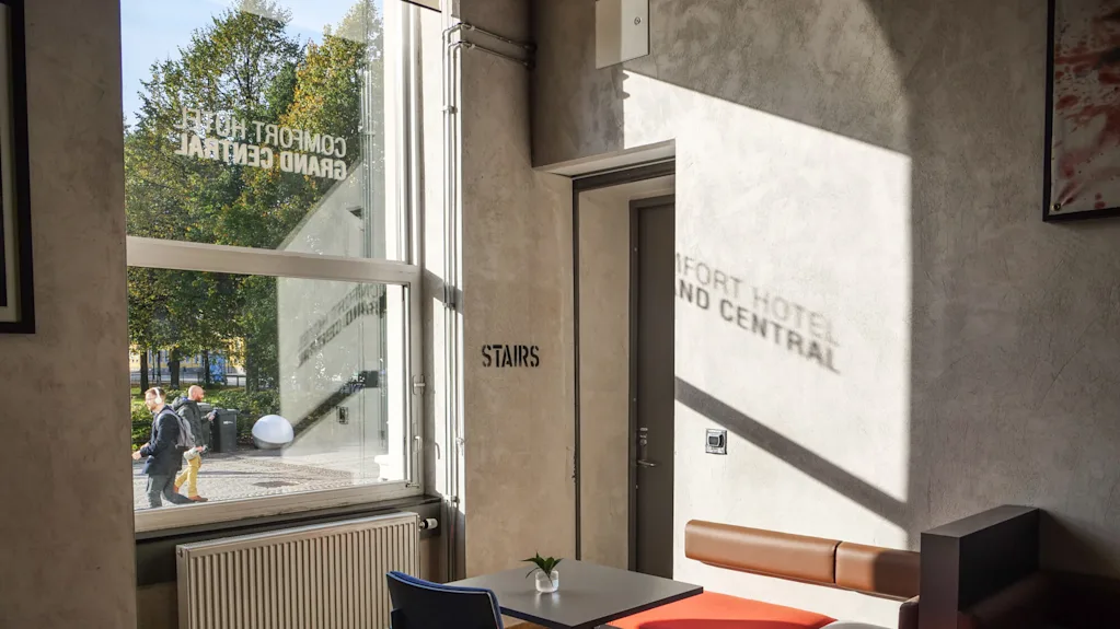 Et vindue viser "COMFORT HOTEL GRAND CENTRAL" og "STAIRS" på en væg og oplyser et moderne siddeområde indeni. Udenfor går to personer forbi træer i sol.