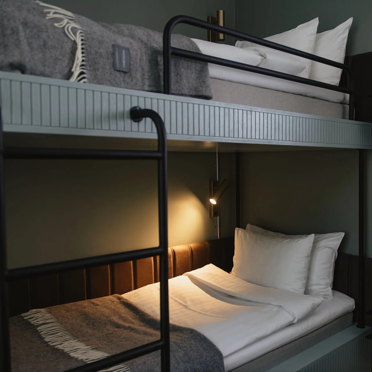 Bunk beds with black metal frames and white bedding are lit by a small wall light. The gray walls create a cozy atmosphere. A gray blanket partially covers the top bed.