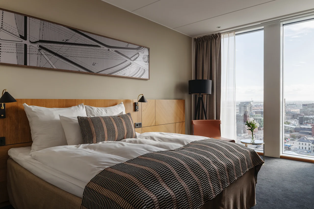 Et moderne hotellrom med en komfortabel seng, et stort vindu som tilbyr byutsikt, og et nattbord med friske blomster.