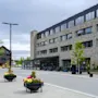 En moderne steinbygning langs en rolig gate, pyntet med fargerike blomsterkasser, flankert av unge trær og et stort banner. Overskyet himmel og gatelykter gir urban stemning.