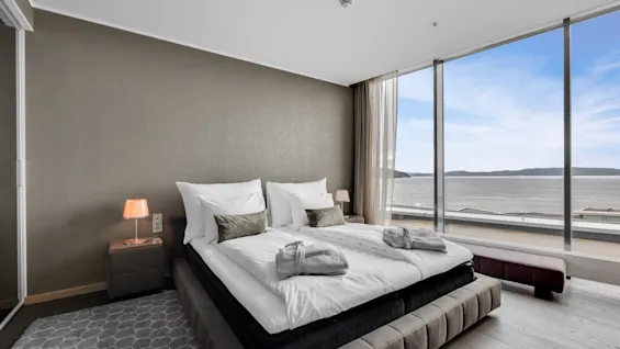 A double bed with white linens and folded robes rests beside a large window overlooking a calm waterfront. The room includes bedside tables with lamps and a gray rug.
