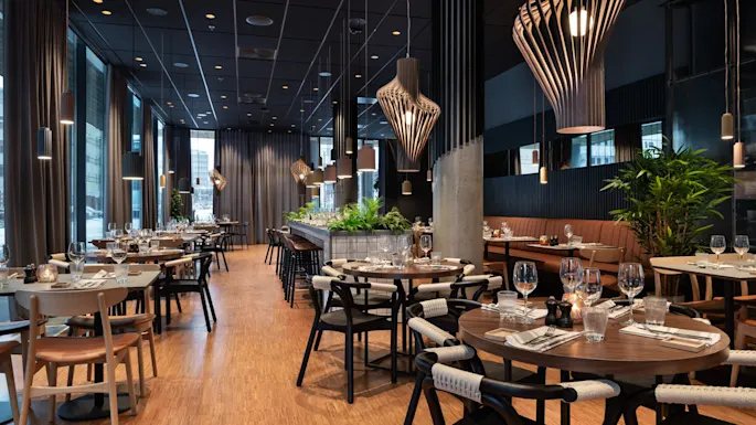 Tables are elegantly set with glassware and cutlery in a modern restaurant, featuring wooden furnishings, stylish pendant lights, and green plants, creating a warm and inviting ambiance.