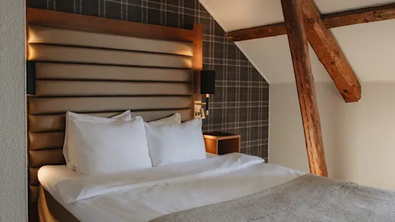 Et hyggeligt hotelværelse med en pænt redt seng, en polstret sengegavl, ternet tapet og rustikke træbjælker.