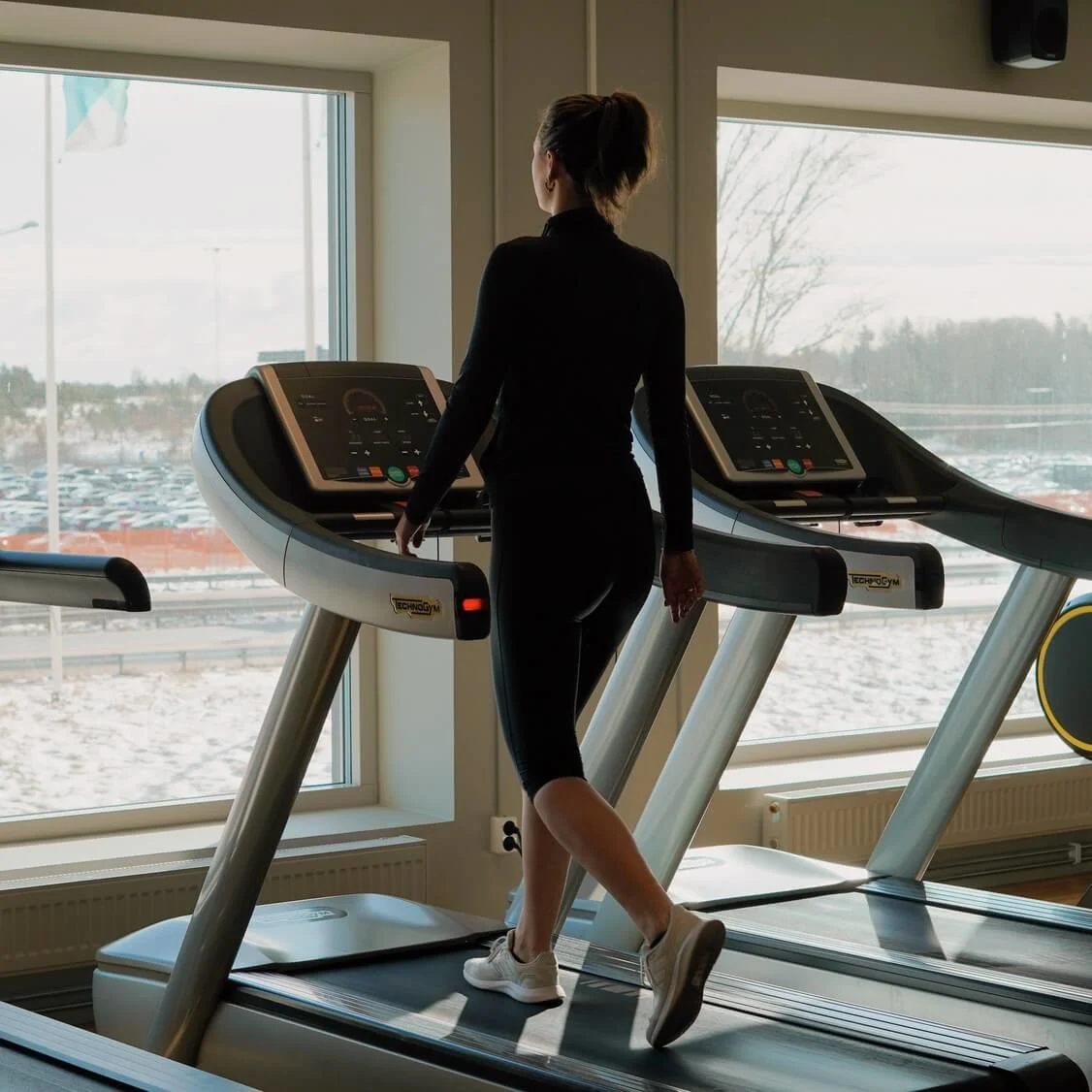 A person walks on a treadmill indoors, gazing outside at a snowy landscape through large windows. The gym is equipped with multiple exercise machines.