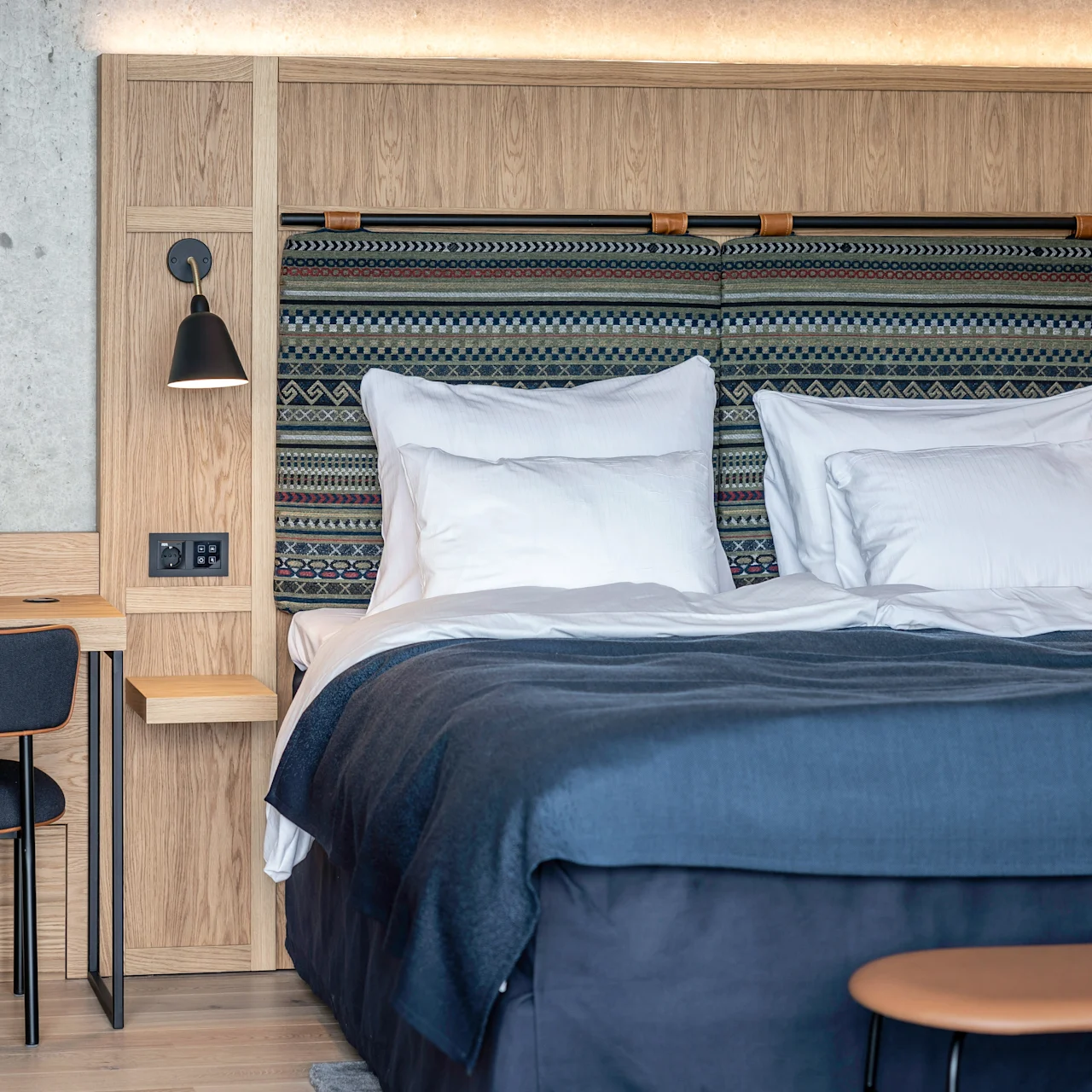 En seng med hvite puter og blå sengesett står mot en vevd mønstret sengegavl. Ved siden av står et treskrivebord med en svart stol under en lampe i et moderne rom.