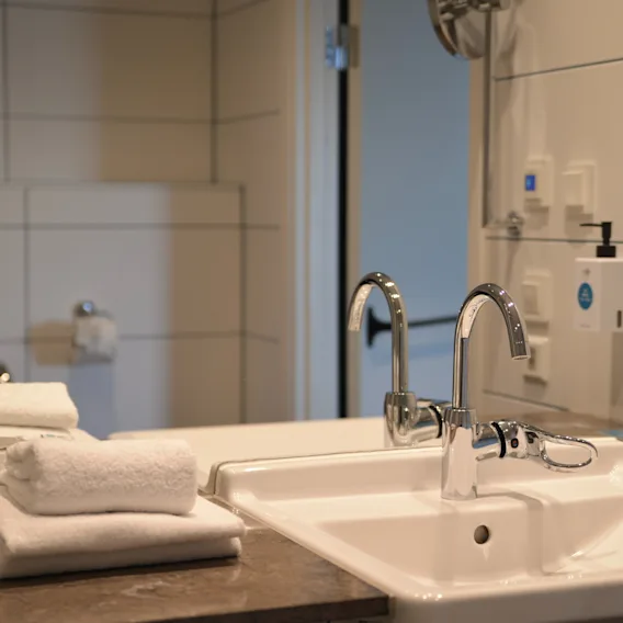 Sink with shiny faucets sits on a stone countertop in a bathroom. White tiles on the walls, a stack of neatly folded towels, and glasses are nearby.