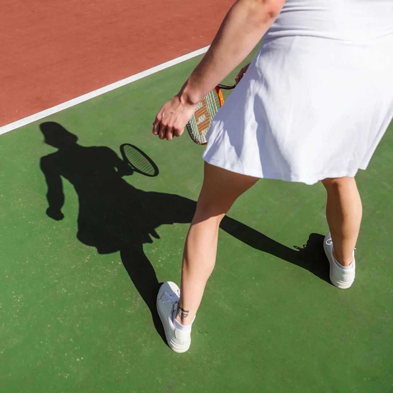 Tennisspiller står med racket, kaster skygge, på en tennisbane med røde og grønne seksjoner.