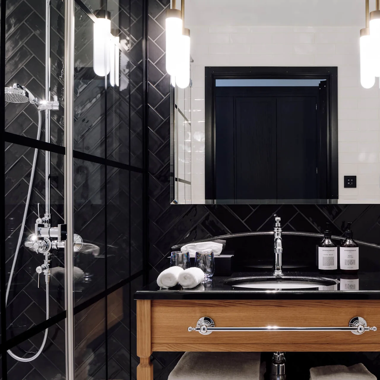 Elegant bathroom with a wooden vanity holding a sink and toiletries. Tiled walls surround with a glass shower visible. Bag labeled "AMERIKALINJEN Hairdryer" hangs nearby. Bright lighting and reflections complete the scene.