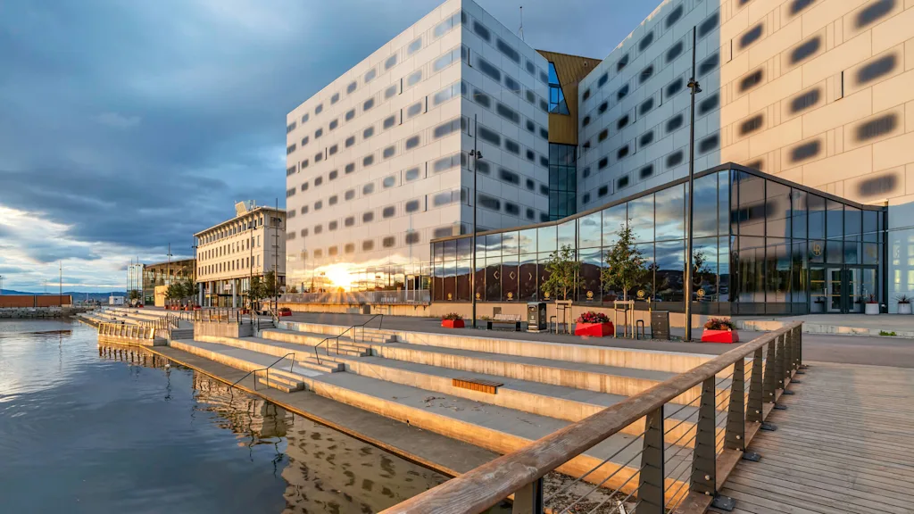 En modern byggnad med reflekterande glasfönster står bredvid en trappad strandpromenad vid vattnet, under en molnig himmel. Röda planteringslådor piffar upp området.