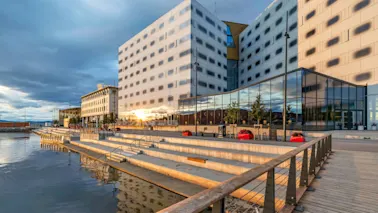 En moderne bygning med reflekterende glassvinduer står ved siden av en trappet promenade ved vannet, under en skyet himmel. Røde plantekasser piffer opp området.