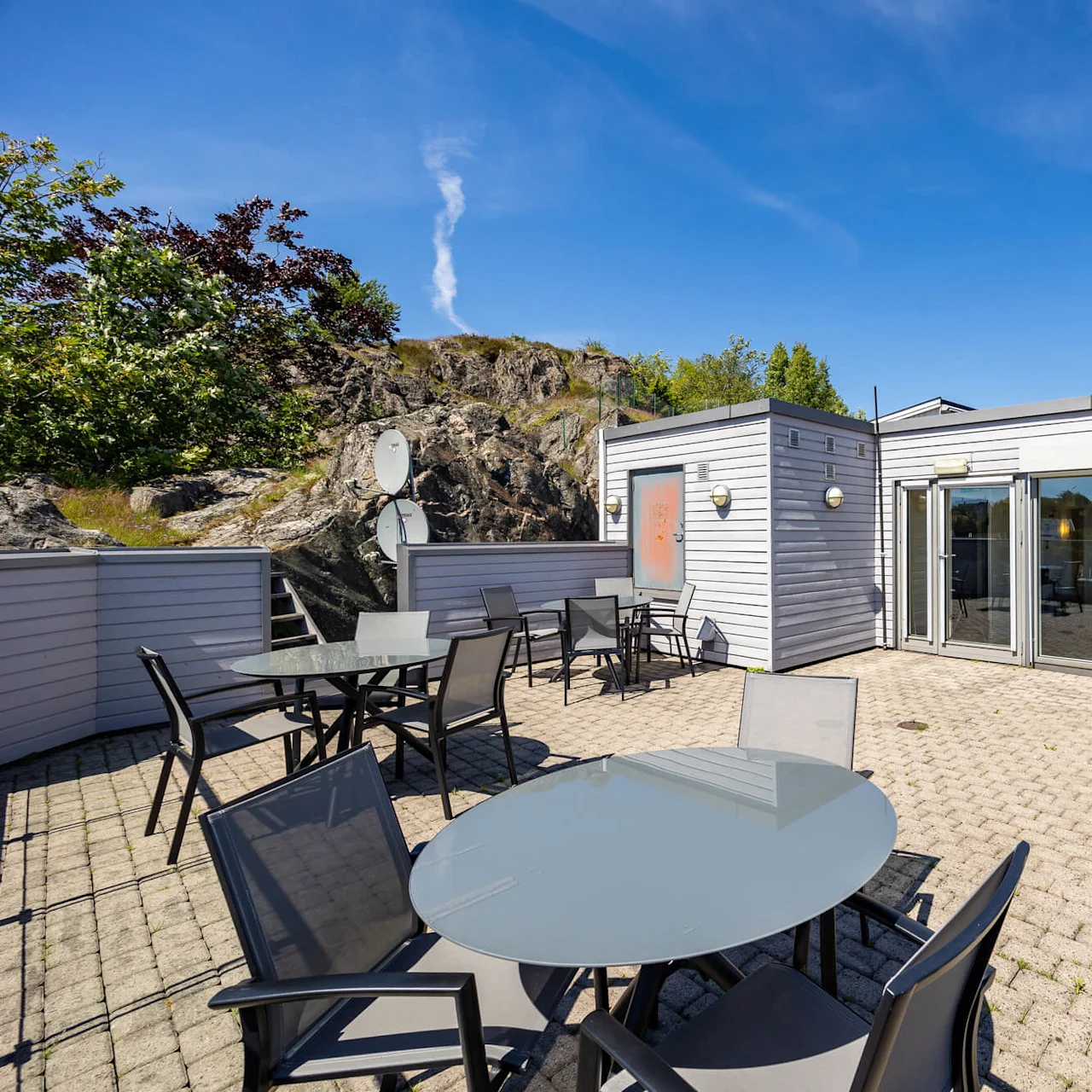 A sunny terrace with tables and chairs outside a modern building with large windows, set against a rocky hill and a clear sky.