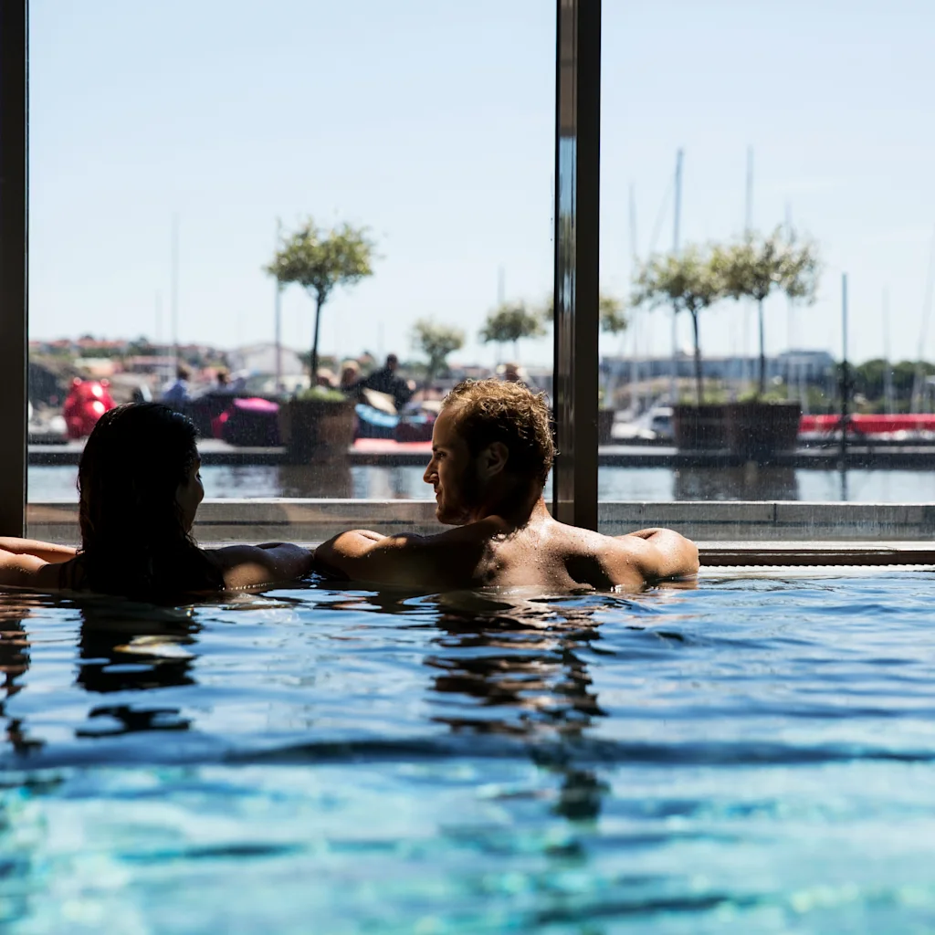 Et par slapper af i en swimmingpool og nyder hinandens selskab. Gennem store vinduer er en marina synlig med fortøjrede både og træer under en klar blå himmel.