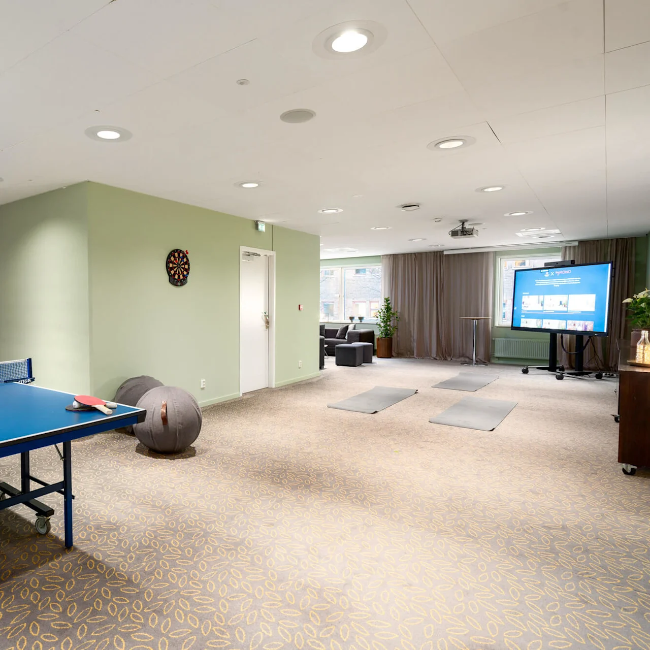Pingisbord med racketar står i ett rymligt rekreationsrum med en darttavla på en ljusgrön vägg, träningsmattor på golvet och en presentationsskärm med texten "Tivoli".