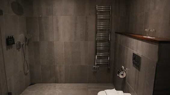 Moderni kylpyhuone tummanharmailla laatoilla, suihkukaappi, lämmitetty pyyheteline ja wc puuhyllyllä.