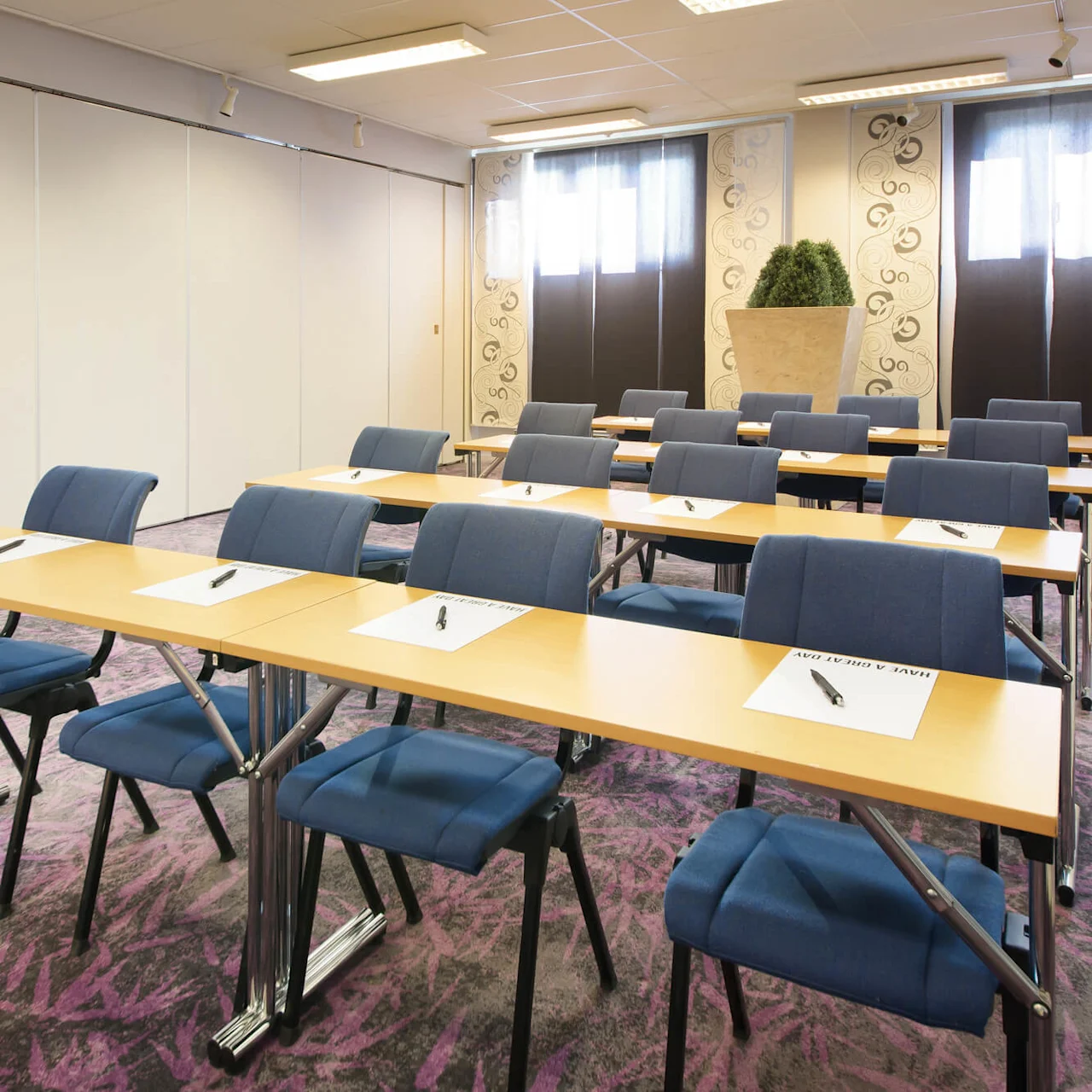 Rader av bord med blå stolar i ett konferensrum, papper och pennor på borden, och lila mattgolv.