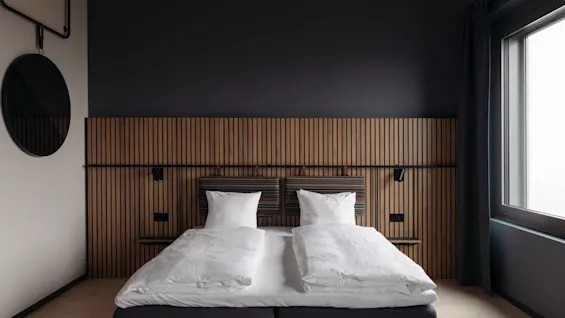 A neatly made bed with white pillows and duvets sits against a wooden panel headboard. A round mirror hangs on the wall, and a window with curtains is on the right.