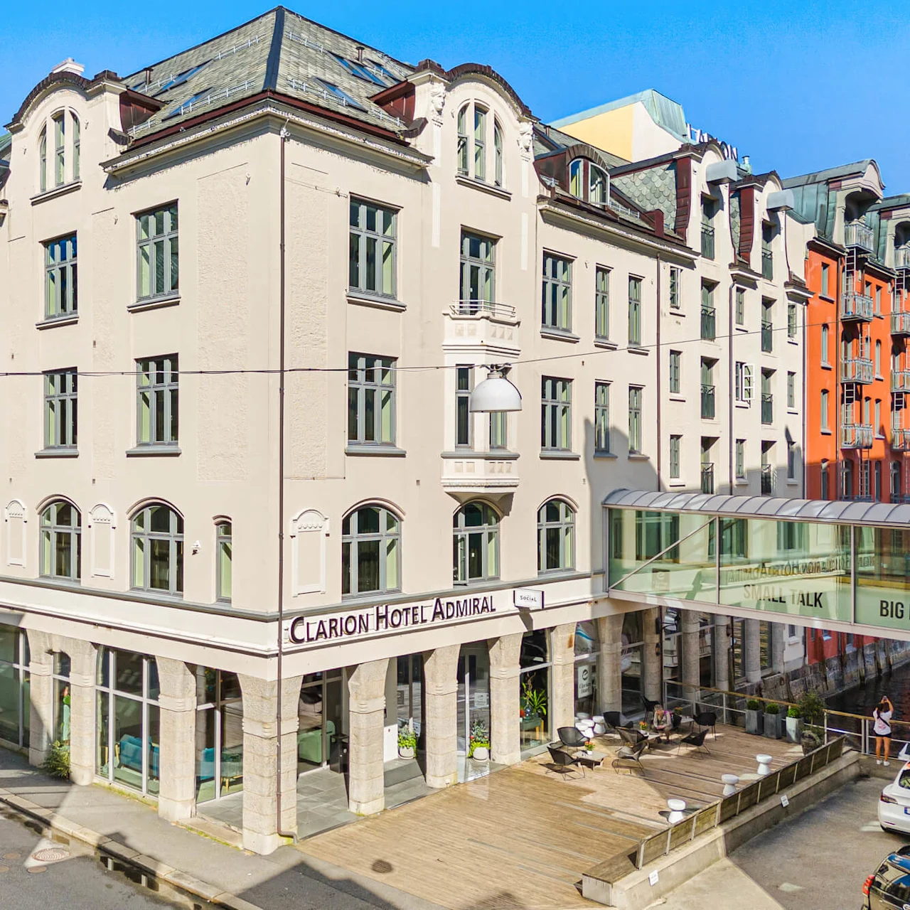 En hjørnebygning mærket "Clarion Hotel Admiral" står højt, med mange vinduer og klassisk design. Hotellet er omgivet af farverige bygninger og en gade med parkerede biler.