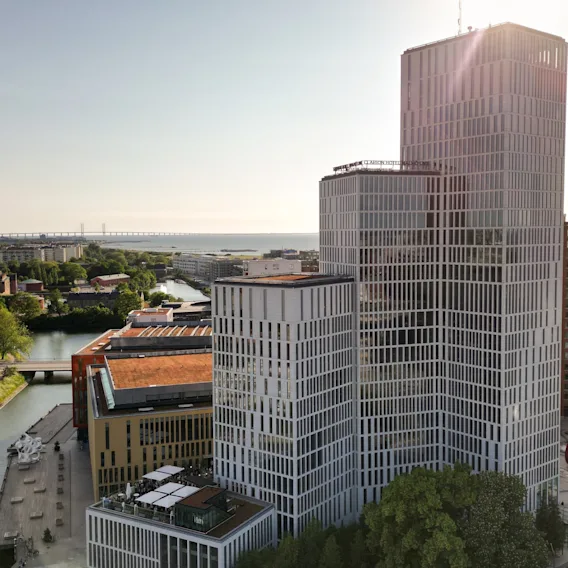 En hög, modern byggnad med rutnätsfasad står vid en kanal i en stadsmiljö med avlägsen utsikt över havet och en bro.
