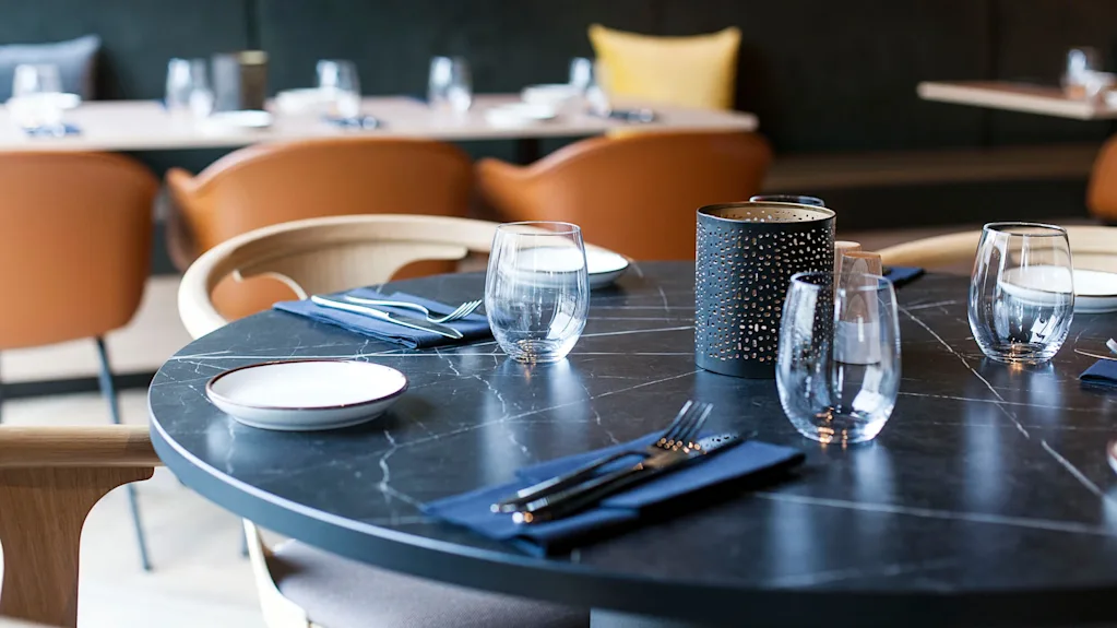Et rundt svart marmorbord har elegant glass, bestikk og et midtstykke i en moderne, mykt opplyst restaurant med brune stoler i bakgrunnen.