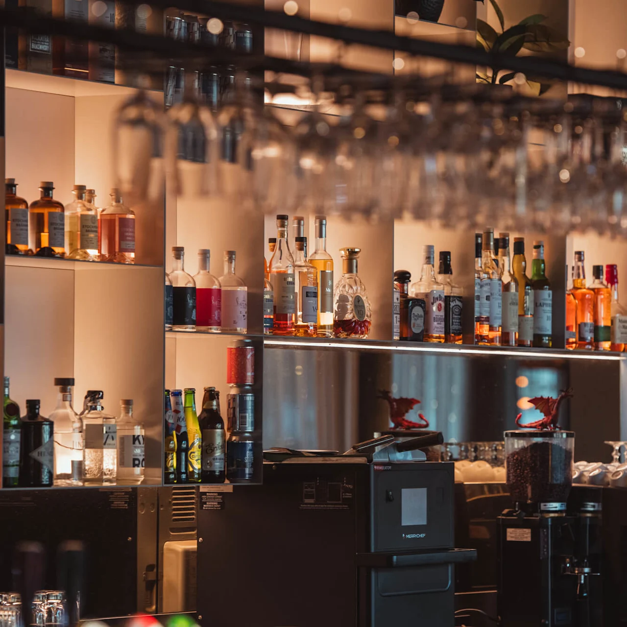 Flaskor med alkohol är prydligt uppställda på hyllor, mjukt upplysta bakifrån. Vinglas hänger upp och ner ovanför. Miljön är en varm, avslappnad bar eller lounge.