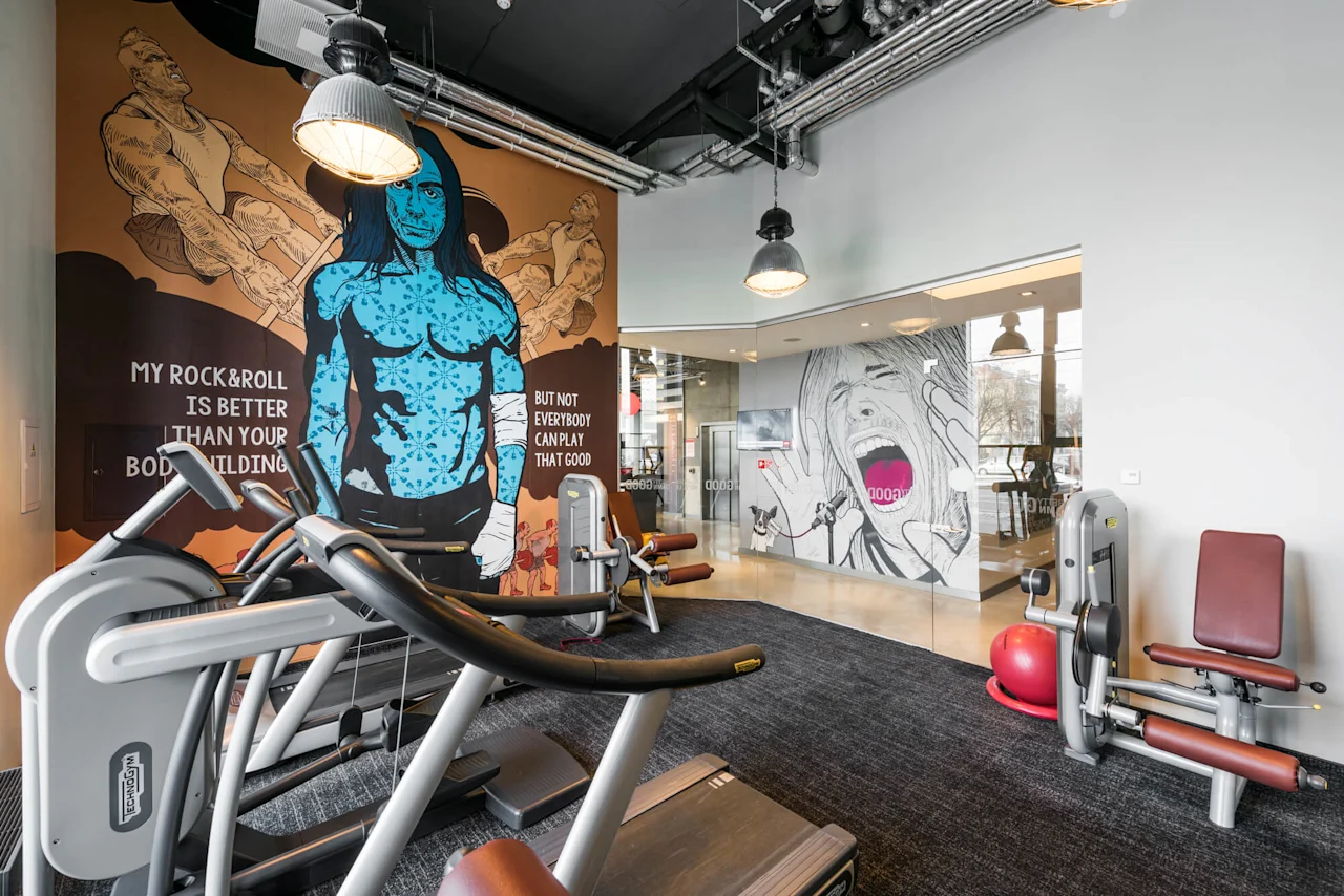 Treadmills and exercise machines in a gym with murals, including a blue figure and people lifting weights. Text: "MY ROCK & ROLL IS BETTER THAN YOUR BODYBUILDING BUT NOT EVERYBODY CAN PLAY THAT GOOD."