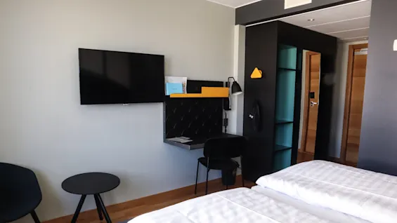 TV monterad på väggen, ovanför ett litet skrivbord med en stol i ett modernt hotellrum; säng, sidobord och öppen dörr synliga. Text på broschyr: "Scandic".