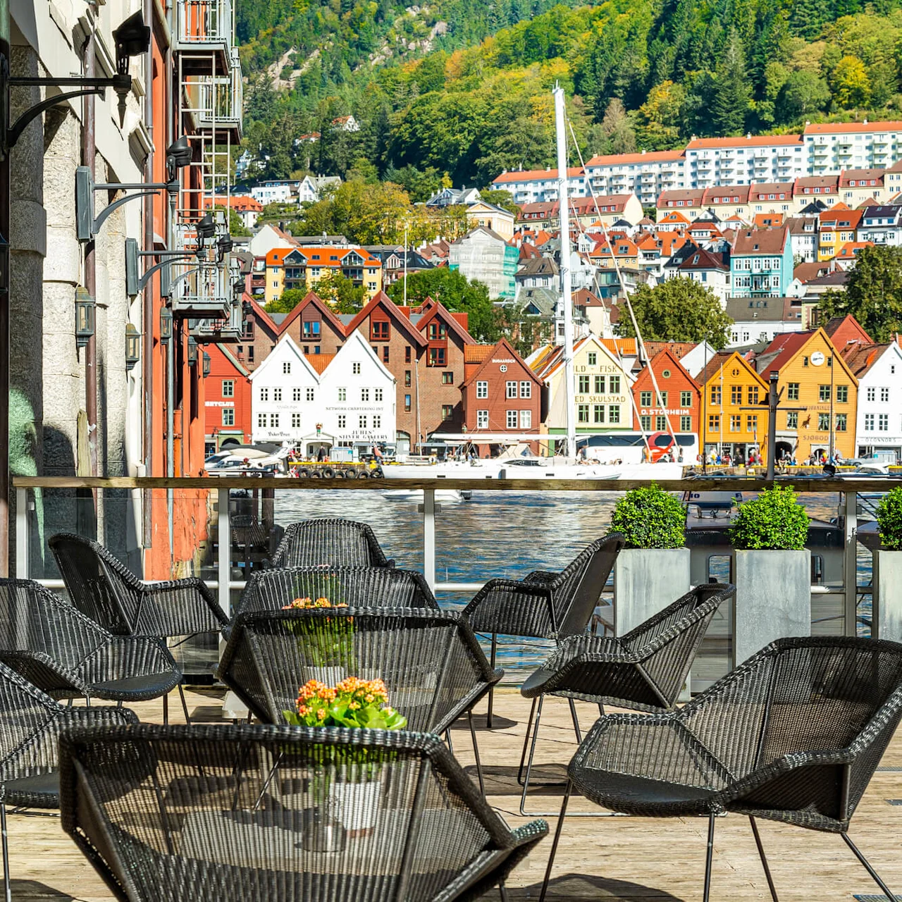 Moderne sorte stole og små borde med blomster på et trægulv med udsigt over farverige bygninger over roligt vand, med en grøn skov og husfyldt bakke i baggrunden.