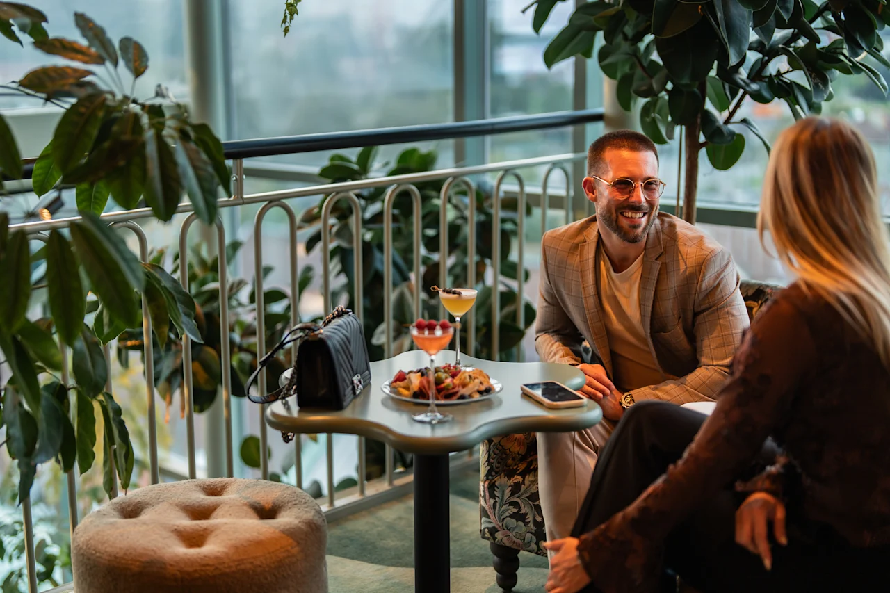 Kaksi ihmistä nauttii juomia ja välipaloja pöydän ääressä valoisassa, kasveilla täytetyssä oleskelutilassa, keskustellen.
