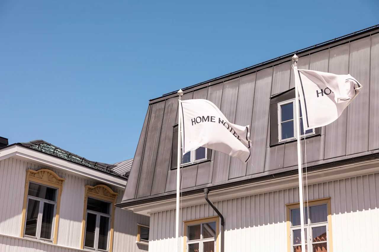 Två flaggor märkta "HOME HOTEL" vajar på stolpar framför en byggnad med sluttande tak och flera fönster under en klarblå himmel.
