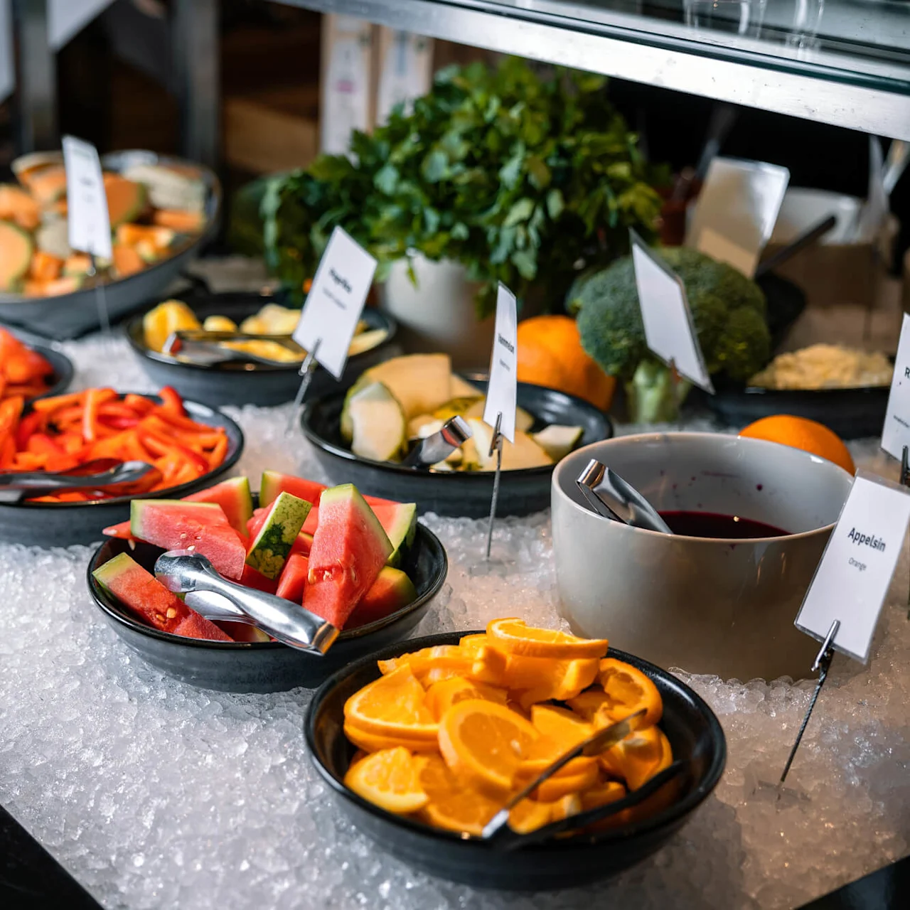 Skåle med skårne frugter og grøntsager, inklusive appelsiner, vandmelon og rød peberfrugt, ligger på en seng af is i en buffetindstilling. Etiketter læses: "Rødbeter," "Appelsin."