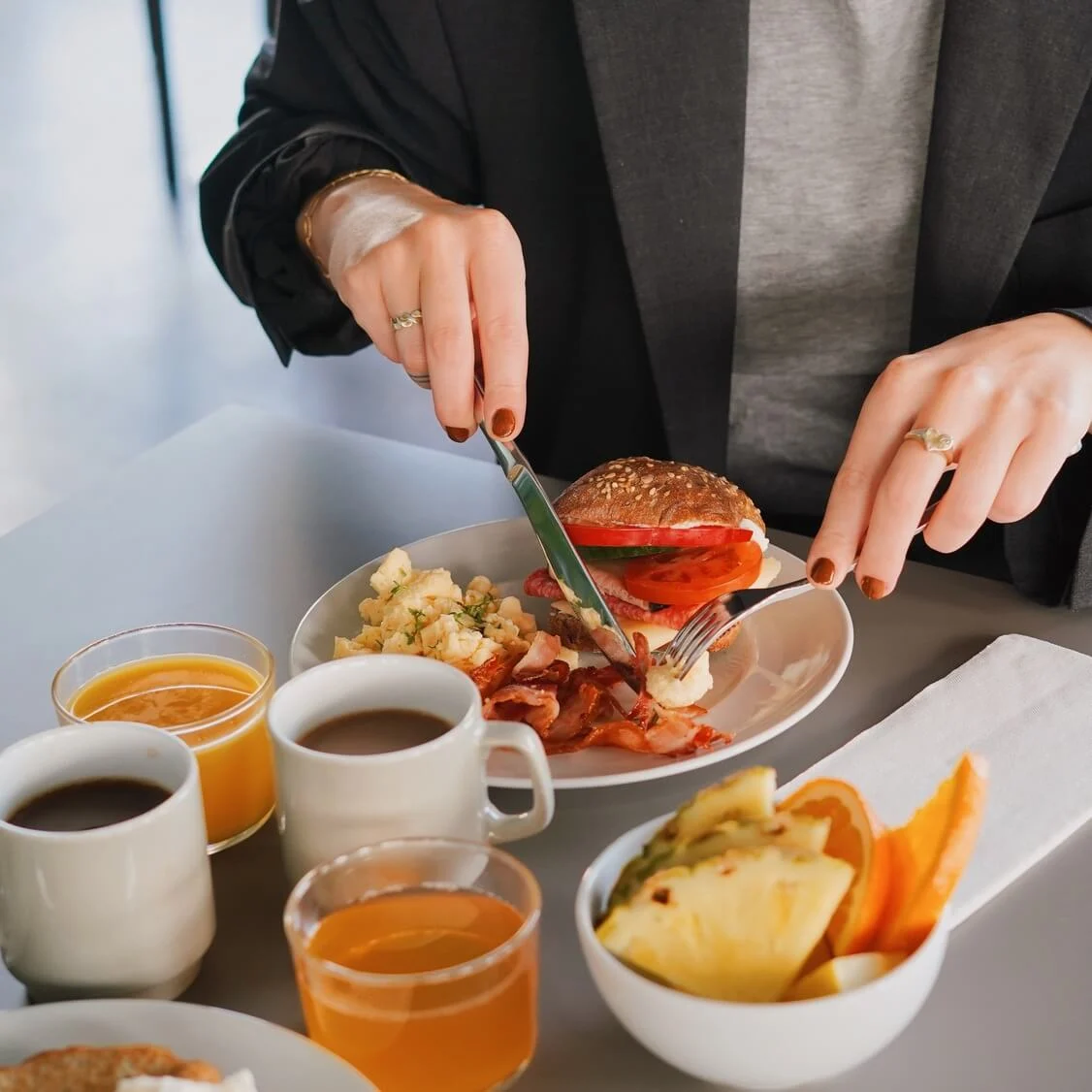 A person wears a dark blazer, cutting a sandwich at a breakfast table with scrambled eggs, bacon, coffee, juice, and fruit in a bright, indoor setting.