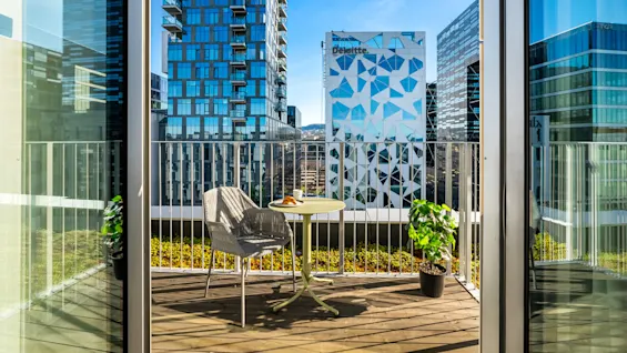 A small balcony with a table, a croissant, and coffee. Surrounded by modern high-rise buildings under a clear blue sky. Visible text: "Deloitte".